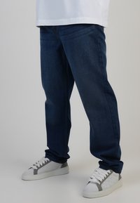 Vaqueros de mezclilla en azul oscuro con un corte recto, que presentan bolsillos frontales y detalles de costura sutiles, combinados con zapatillas blancas y acentos grises.