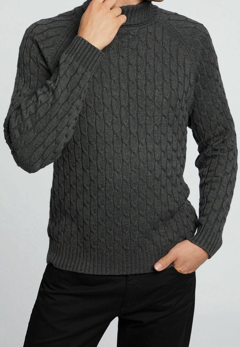 Musta harmaa cable-knit-neulepusero korkealla kauluksella, jossa on tekstuurikuvioita ja ribattuja hihoja. Yhdistetty mustiin housuihin, käsi levähtää lantiolla.
