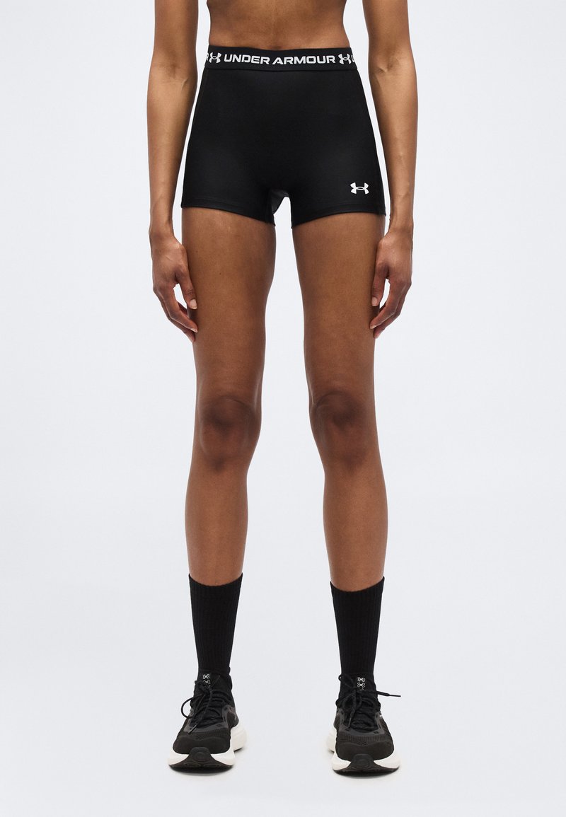 Άτομο που φοράει μαύρα αθλητικά σορτς Under Armour, μαύρες κάλτσες μέσης και μαύρα παπούτσια για τρέξιμο, στέκεται όρθιο μπροστά σε απλό λευκό φόντο.