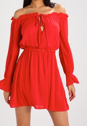 Femme portant une robe rouge vif décolletée aux épaules, manches longues, poignets à volants et taille cintrée avec un détail de lien à l'avant.