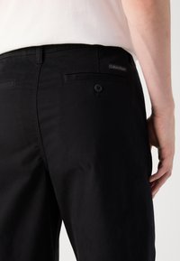 Black cotton trousers with a smooth texture. Features a back pocket with a button and a small logo tag on the waistband.