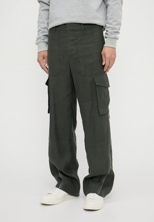 Cargo pants in dark green, featuring a loose fit, two side pockets, and zippers near the hem. Made from a textured fabric. White sneakers.