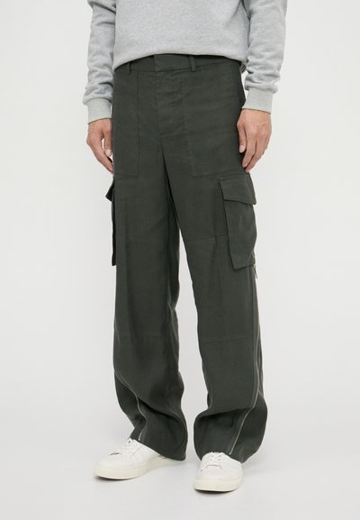 Pantalons cargo de couleur vert foncé, avec une coupe ample, deux poches latérales et des zips près de l'ourlet. Fabriqués dans un tissu texturé. Baskets blanches.