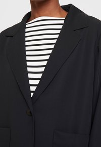 Blazer noir à col cranté avec fermeture par un seul bouton, porté par-dessus un haut rayé blanc et noir. Tissu lisse, coupe ajustée.