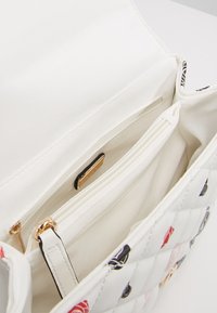 Bílá prošívaná kabelka uvnitř s dvojitými zipovými oddíly a hladkým textilním podšívkou. Obsahuje lesklé zlaté kovové detaily.
