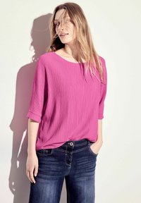 Top rosa con texture, dal collo rotondo e maniche a tre quarti, abbinato a jeans blu scuro con cuciture a vista e una tasca frontale.