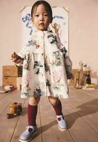 Weißes Kleid mit bunten Tier- und Pflanzenmotiven, Rüschensleeves, kombiniert mit roten kniehohen Socken und blauen Schuhen. Holzspielzeug im Hintergrund.
