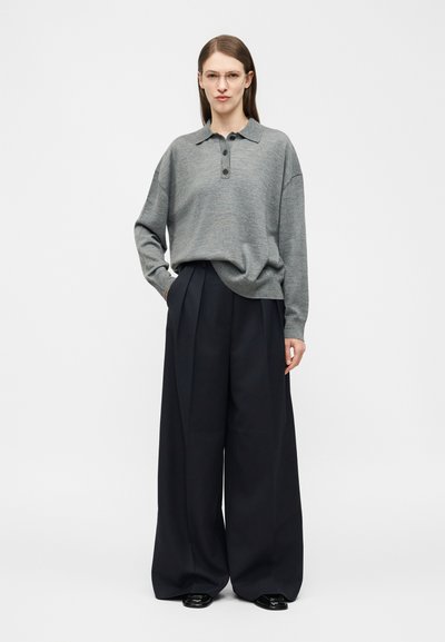 Kobieta nosząca przezroczyste okulary, szary sweter polo z długim rękawem oraz czarne spodnie z szerokimi nogawkami, stojąca z jedną ręką w kieszeni.