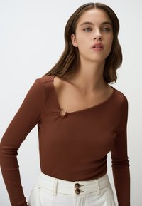 Braun gestreiftes Langarmoberteil mit asymmetrischem Kragen und einem dekorativen Schlaufenakzent. Das Modell trägt es mit hoch geschnittenen weißen Hosen.