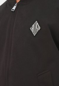 Must musta jope, millel on sile tekstuur, varustatud tõmbluku sulgemisega ja vasakul rinnal rombikujuline tikitud logo.