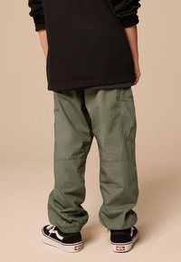 Pantalon cargo vert avec poignets élastiques, poche latérale, et coupe décontractée, associé à des baskets noires avec lacets blancs et détail logo.