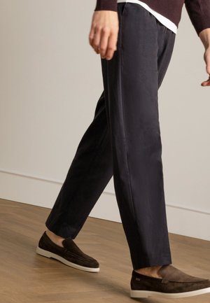 Marineblauwe broek met een rechte snit, gemaakt van zachte stof, met een soepele textuur. Gecombineerd met donkerbruine instappers op een houten vloer.