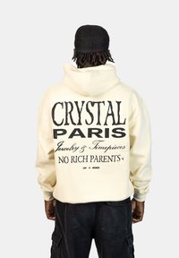 Crèmekleurige hoodie met grote zwarte tekst op de achterkant: "CRYSTAL PARIS Sieraden & Horloges GEEN RICHTE OUDERS." Eenvoudig ontwerp, zachte textuur.