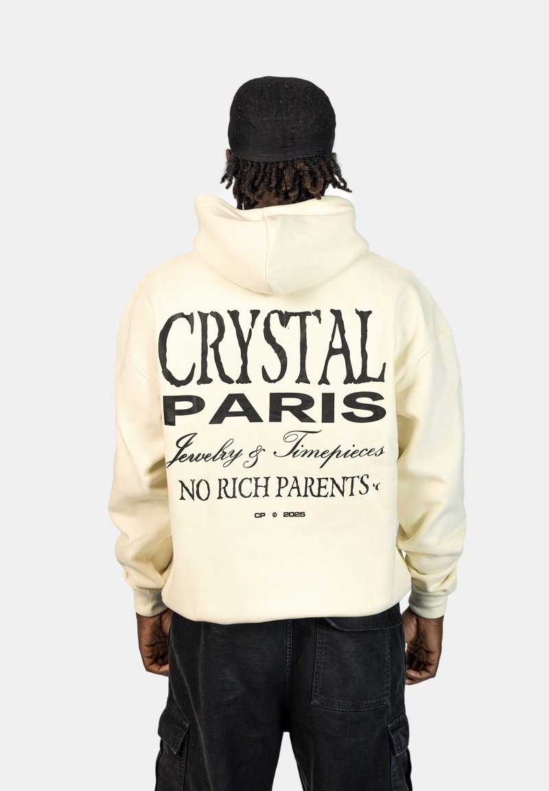Crèmekleurige hoodie met grote zwarte tekst op de achterkant: "CRYSTAL PARIS Sieraden & Horloges GEEN RICHTE OUDERS." Eenvoudig ontwerp, zachte textuur.