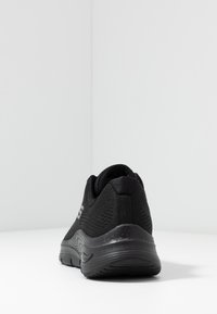 Zapatilla deportiva negra con parte superior de malla texturizada, cuello acolchado y suela acolchada que presenta una curva pronunciada y un detalle sutil.