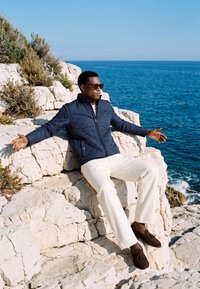 Homme portant des lunettes de soleil, une veste marine, un pantalon blanc et des chaussures marron, assis bras écartés sur une falaise rocheuse blanche face à l'océan bleu.