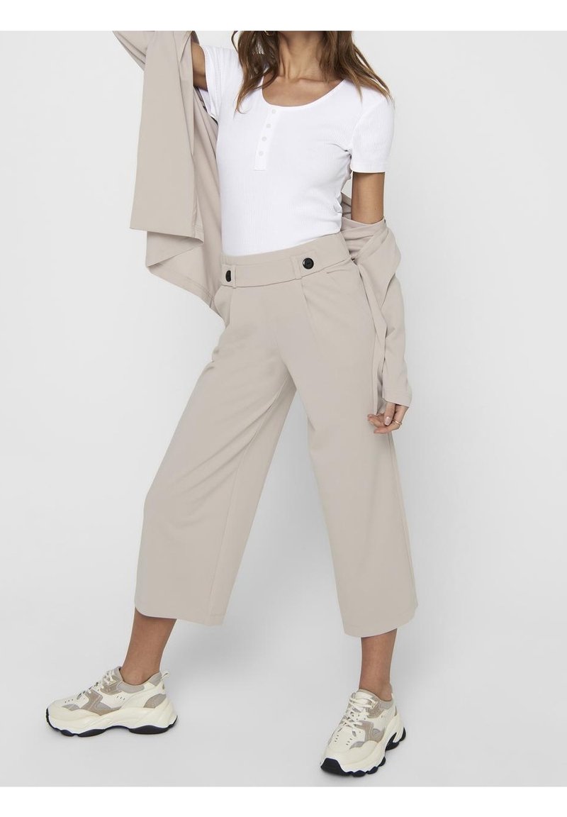 Femme portant un pantalon beige large et raccourci, une chemise blanche à manches courtes, une veste beige drapée sur les épaules et des baskets blanches épaisses.