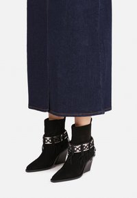 Botines de ante negro hasta el tobillo con punta afilada, detalles decorativos en plata y borlas. Combinados con una falda de mezclilla oscura.