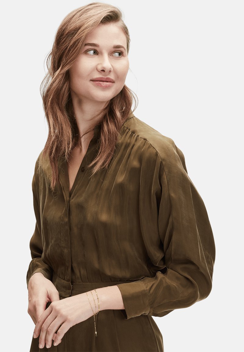 Olivgrön silkesblus med knapplist, plisserade detaljer och långa ärmar. Accessoarer inkluderar lager av guldarmband på handleden.