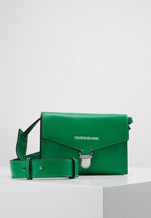 Grøn læder crossbody-taske fra Calvin Klein Jeans med sølvspænde og justerbar rem, vist på hvid overflade mod grå baggrund.