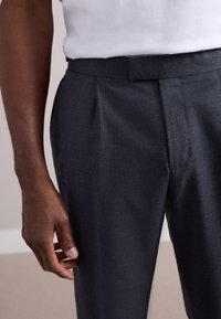 Grey tailored pants with a textured finish, pleats at the front, slim fit, flat waistband, and hand resting on the side.