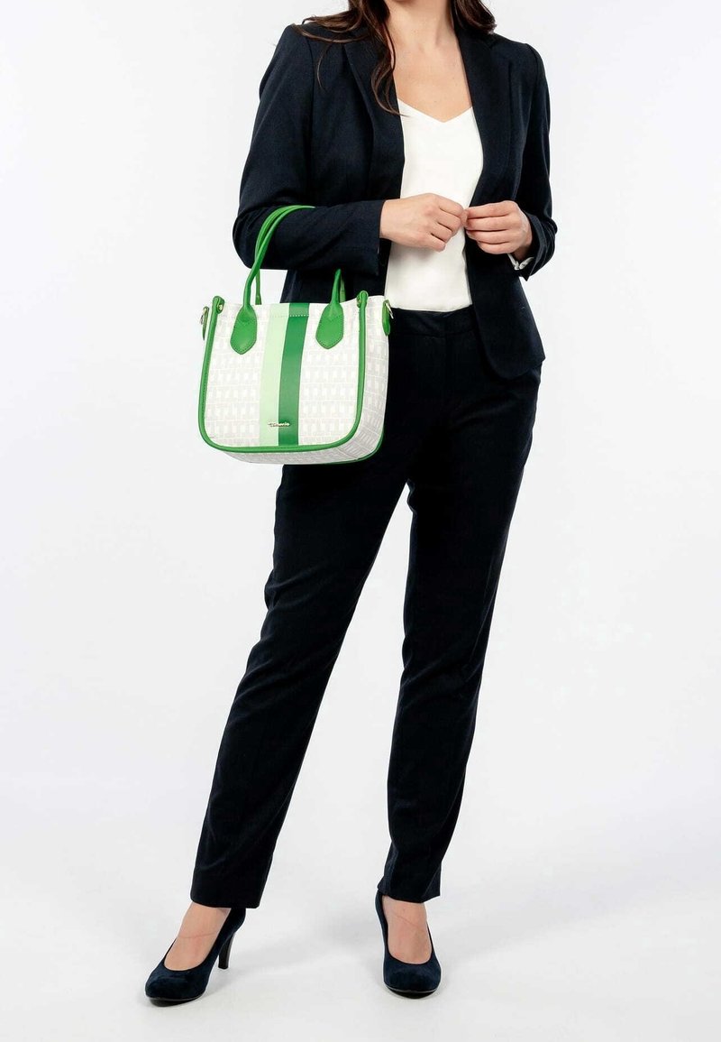 Femme en costume noir et blouse blanche tenant un sac à main blanc avec poignées vertes et bande, debout devant un fond blanc uni.