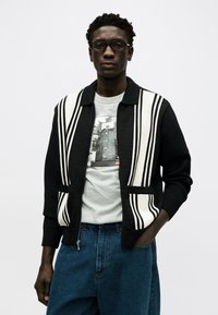 Άνδρας που φοράει μαύρη και λευκή ριγέ ζακέτα, γκρι μπλουζάκι με στάμπα, μπλε τζιν και γυαλιά, στέκεται με το ένα χέρι στην τσέπη.