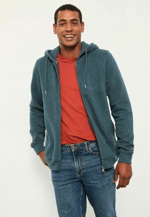 Hooded zip-up sweatshirt in dark teal, made of soft fabric. Layered over a red shirt, paired with blue jeans. Features drawstrings and cuffs.