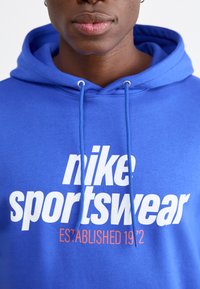 Felpa blu con testo bianco "nike sportswear" e dettaglio arancione "ESTABLISHED 1972". Presenta un cappuccio con coulisse e una morbida texture di tessuto.