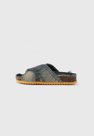 Denim slide sandal with crisscross straps, frayed edges, and a textured brown sole featuring a zigzag pattern. Color: blue with brown accents.