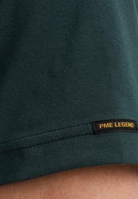 Tamnozeleni majica s glatkom teksturom i uskim krojem. Mali crni etiketa s natpisom "PME LEGEND" u žutoj boji ušivena je blizu donjeg ruba.