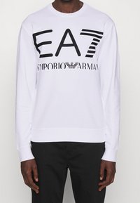 Vit sweatshirt i bomull med svart grafisk text "EA7 EMPORIO ARMANI" i olika stilar. Ribbad mudd och nederkant.