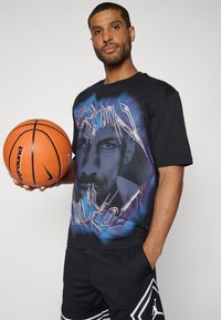 Camiseta negra con un gran estampado gráfico que presenta una cara y acentos azules. El modelo sostiene un balón de baloncesto naranja, vistiendo pantalones cortos negros.