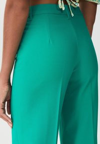 Gros plan sur une personne portant un pantalon turquoise taille haute avec passants de ceinture, montrant une partie de sa main et de sa taille.