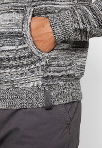 Suéter de punto con rayas grises y negras, bolsillo tipo canguro, puños acanalados y una etiqueta tejida en el dobladillo. Textura suave.