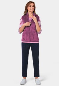Lila Fleece-Weste mit hohem Kragen, gestreiftem Langarmshirt, navy Hosen und weißen Sneakers. Verfügt über einen Reißverschluss und eine weiche Textur.
