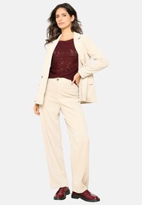Bež žametni blazer in ujemajoče se široke hlače nad maroon pleteno majico, obutev v burgundski barvi; gladka tekstura in prilagojen kroj.