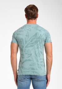 Šviesiai mėlyna trumpomis rankovėmis marškinėliai, puikuojantys tekstūruotu lapų raštu. Audinys atrodo minkštas su subtiliu blizgesiu, kuris puikiai dera su mėlynais džinsais.