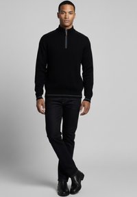 Suéter negro acanalado con cremallera y cuello alto, combinado con vaqueros oscuros y botas negras, con un sutil ribete de contraste en el cuello.