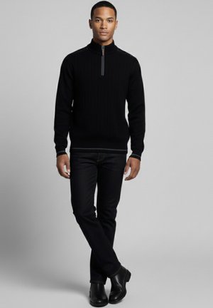 Suéter negro acanalado con cremallera y cuello alto, combinado con vaqueros oscuros y botas negras, con un sutil ribete de contraste en el cuello.