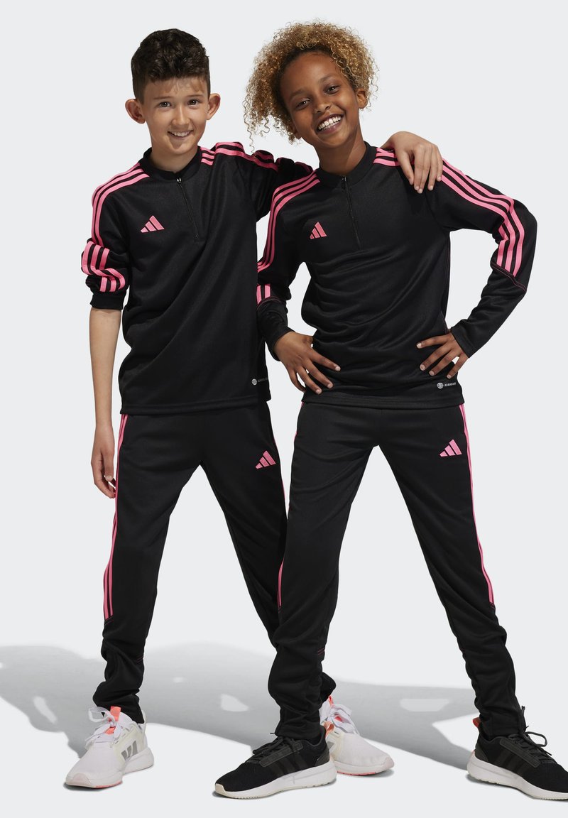 Schwarze sportliche Trainingsanzüge mit pinken Streifen, mit Reißverschlusskragen, weicher Textur, mit dem Adidas-Logo auf der Brust und den Hosen, getragen von zwei Kindern.