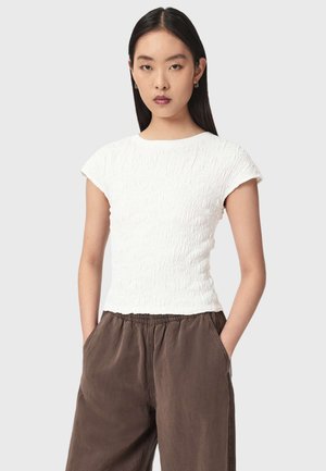 Junge Frau mit langen dunklen Haaren trägt ein strukturiertes weißes Kurzarmshirt und lockere braune Hosen, die Hände in den Taschen, stehend vor einfarbigem Hintergrund.
