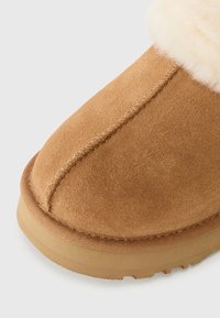 Brune semskede støvler med en rund tå, som har en fluffy kremfarget krage og en gummisåle. Synlig søm langs sømmene.