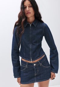 Giacca di denim con design corto e chiusura con zip, abbinata a una mini gonna in denim coordinata. Completata con cuciture a contrasto e tasche standard.