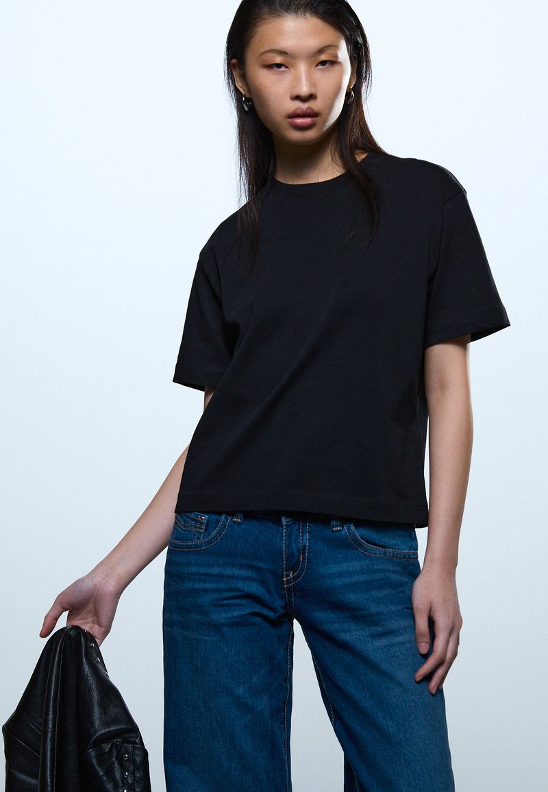 Νεαρή γυναίκα που φοράει μαύρο μπλουζάκι και μπλε τζιν, κρατώντας στο αριστερό χέρι της ένα διπλωμένο μαύρο δερμάτινο μπουφάν.