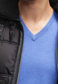 Blau gestrickter V-Ausschnitt-Pullover kombiniert mit einer schwarzen gepolsterten Jacke, die einen Kragen aus Kunstschafswolle und metallische Hardware-Details aufweist.