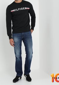 Svart sweatshirt med texten "HILFIGER" och röda/vita ränder, kombinerad med blå denimjeans och svarta sneakers. Enkel design och avslappnad passform.