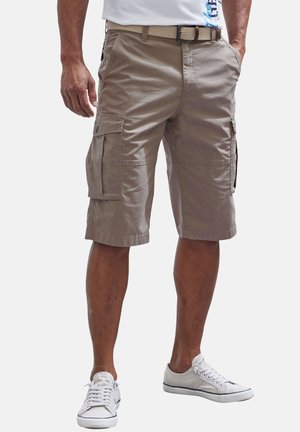 Man wearing beige cargo shorts, white belt, white casual sneakers, standing with one hand in pocket against plain background.