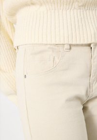 Suéter de punto acanalado color crema con un cuello alto, combinado con pantalones de pana beige claro que cuentan con bolsillos delanteros y costuras sutiles.