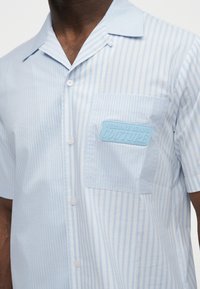 Homme portant une chemise à manches courtes boutonnée à rayures verticales bleu clair et blanc avec une poche comportant l'inscription « Versace Jeans Couture ».